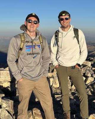 Joe, on the left in a grey hoodie, and a buddy standing on a rocky summit in low sun, both smiling in sunglasses, beanies with headlamps, and daypacks, with hazy mountain ridges behind them.