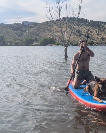 Me kneeling on a red and blue paddleboard on a lake, holding a paddle, with Sasha lying on the front of the board. Dry hills and a bare tree standing in the water behind.